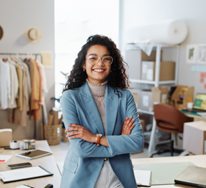 Eine Schneiderin steht in ihrer Werkstatt. Sie hat die Arme verschränkt und blickt in die Kamera.