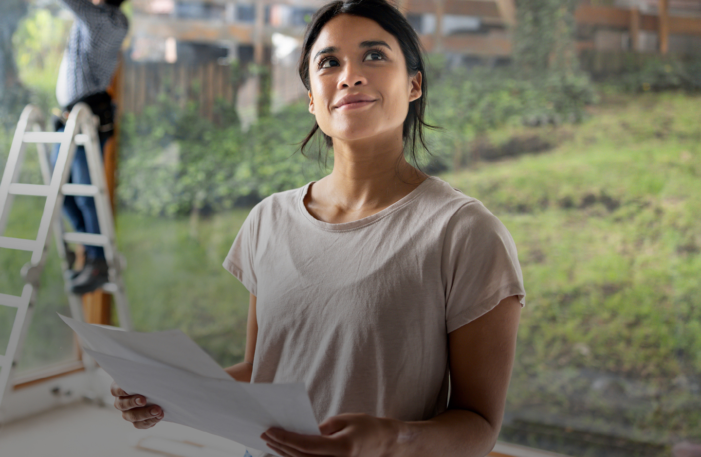 Eine junge Frau steht in ihrem neuen Haus, das gerade gebaut wird. Sie hat einen Bauplan in der Hand.